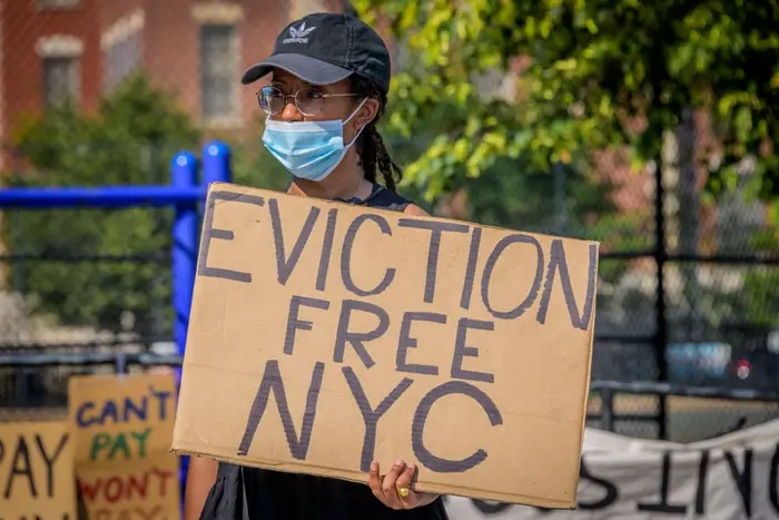 A protestor holds a cardboard sign saying "eviction free NYC" while wearing a blue face mask